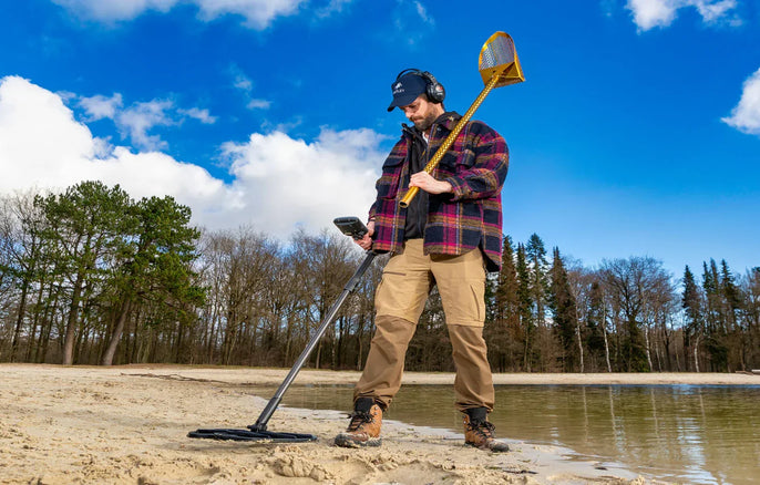 Détectoriste équipé d’un détecteur de métaux sur une plage de sable au bord de l’eau avec pelle de récupération pour la détection de loisirs.